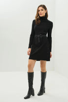 Woman wearing a black dress with a belt and black knee-high boots on a white background